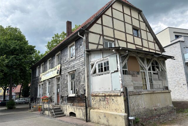Das Foto zeigt ein älteres Fachwerkhaus unter einem bewölkten Himmel. Teile des Hauses sind mit Schindeln verkleidet, während ein Anbau deutliche Gebrauchsspuren und Graffiti aufweist.