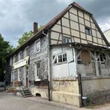 Das Foto zeigt ein älteres Fachwerkhaus unter einem bewölkten Himmel. Teile des Hauses sind mit Schindeln verkleidet, während ein Anbau deutliche Gebrauchsspuren und Graffiti aufweist.