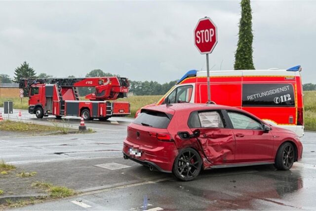 Ein beschädigtes rotes Auto steht auf einer Kreuzung. Dahinter stehen ein Rettungs- und ein Feuerwehrfahrzeug.