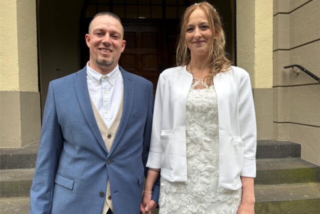 Ein frisch vermähltes Paar posiert Händchen haltend vor einem Gebäude. Die Frau trägt ein weißes Kleid mit Spitze und eine kurze weiße Jacke, während der Mann einen blauen Anzug mit beiger Weste trägt.