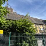 Ein Gebäude steht hinter einem Zaun. Davor stehen Bäume und Sträuche.