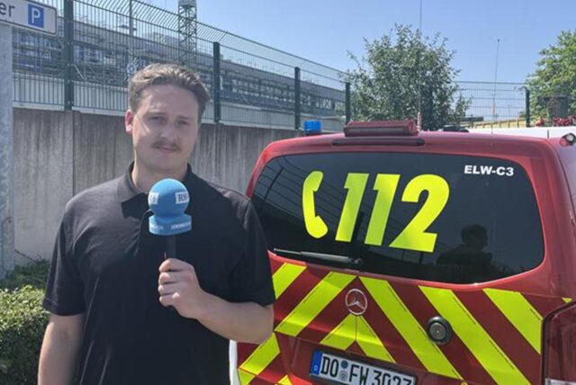 RN-Reporter Tim Hübbertz steht mit einem Mikrofon in der Hand am REWE-Zentrallager in Dortmund.