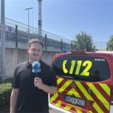 RN-Reporter Tim Hübbertz steht mit einem Mikrofon in der Hand am REWE-Zentrallager in Dortmund.