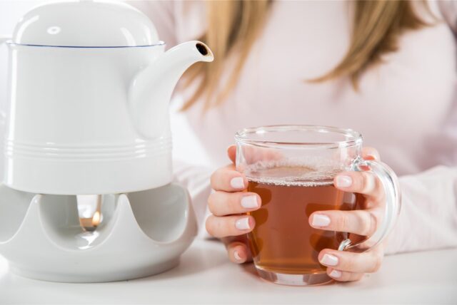 Eine Frau sitzt an einem Tisch und hält eine Tasse Tee in den Händen, die neben einer Teekanne steht.