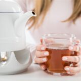 Eine Frau sitzt an einem Tisch und hält eine Tasse Tee in den Händen, die neben einer Teekanne steht.