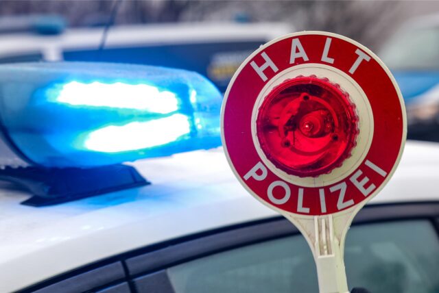 Ein Polizist hält eine Polizeikelle für eine Kontrolle vor einem Einsatzfahrzeug hoch.