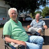 Zwei Personen sitzen im Freien auf Klappstühlen. Im Hintergrund sind Wohnhäuser und geparkte Autos zu sehen.