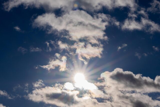 Das Bild zeigt einen strahlend blauen Himmel, teilweise verdeckt von weißen Wolken. Die Sonne scheint hell und durchbricht die Wolkendecke.