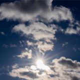 Das Bild zeigt einen strahlend blauen Himmel, teilweise verdeckt von weißen Wolken. Die Sonne scheint hell und durchbricht die Wolkendecke.