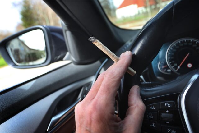 Ein Mann sitzt mit einem Joint zwischen den Fingern am Steuer eines Autos. (Symbolbild)