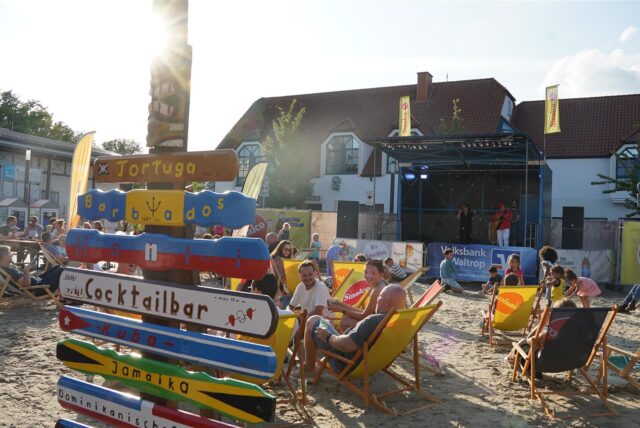 Menschen mit Liegestühlen auf einer Sandfläche auf einem Marktplatz.