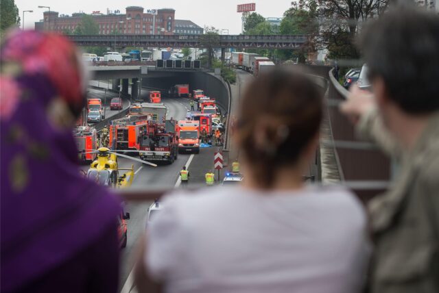 Menschen stehen auf einer Brücke und blicken runter auf einen Verkehrsunfall mit Rettungskräften und Rettungshubschrauber.