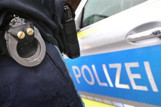 An dem Gürtel eines Polizisten sind Handschellen und eine Pistole zu sehen, im Hintergrund steht sein Streifenwagen.