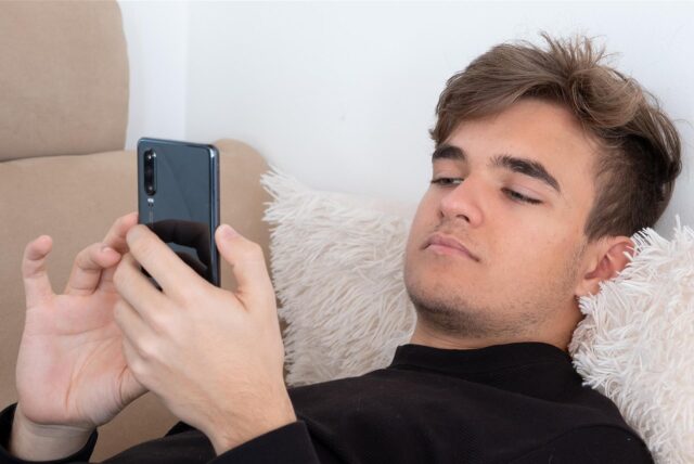 Junger Mann surft am Handy, auf dem Sofa liegend.