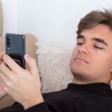 Junger Mann surft am Handy, auf dem Sofa liegend.