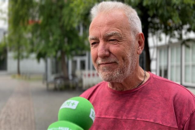 Ein Mann mit grauem Haar wird interviewt. Zu sehen sind grüne Mikrofone und ein rotes Hemd.