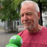 Ein Mann mit grauem Haar wird interviewt. Zu sehen sind grüne Mikrofone und ein rotes Hemd.