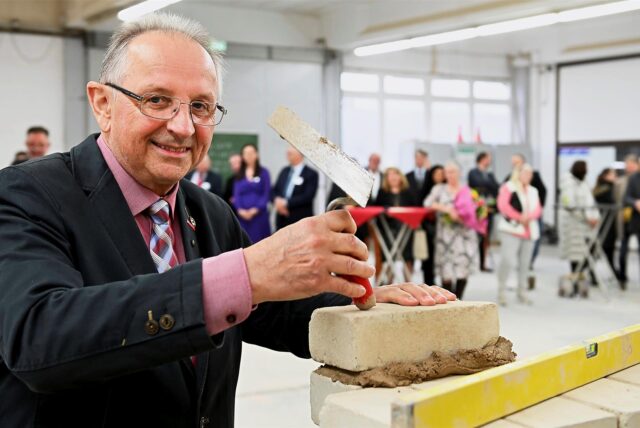 Heinrich Kinzler hält eine Maurerkelle in der Hand und setzt einen Ziegelstein auf eine bereits gemauerte Reihe. Im Hintergrund sind weitere Personen zu sehen, die dem Ereignis beiwohnen.