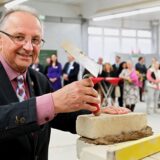 Heinrich Kinzler hält eine Maurerkelle in der Hand und setzt einen Ziegelstein auf eine bereits gemauerte Reihe. Im Hintergrund sind weitere Personen zu sehen, die dem Ereignis beiwohnen.