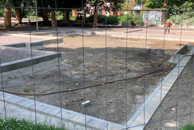 Auf dem Bild ist eine Baustelle hinter einem Bauzaun zu sehen. Im Hintergrund sind ein Bagger und eine Person mit einer Harke zu erkennen.
