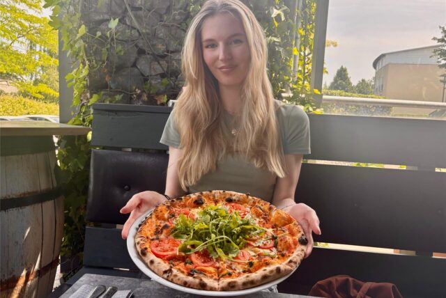Eine junge Frau präsentiert eine Pizza auf einem Teller. Die Pizza ist mit Tomaten und Rucola belegt.