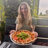 Eine junge Frau präsentiert eine Pizza auf einem Teller. Die Pizza ist mit Tomaten und Rucola belegt.