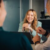 Eine Frau sitzt mit einem Lächeln an einem Tisch mit einem Laptop, während zwei weitere Personen sich mit ihr unterhalten.