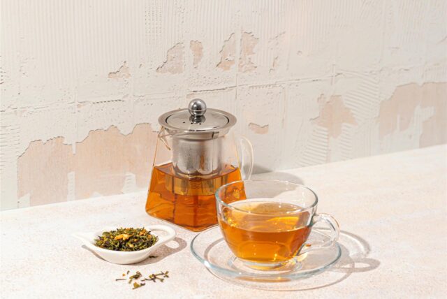 Eine Teekanne aus Glas, gefüllt mit Tee, neben einer gefüllten Teetasse aus Glas auf einem Tisch.