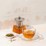 Eine Teekanne aus Glas, gefüllt mit Tee, neben einer gefüllten Teetasse aus Glas auf einem Tisch.