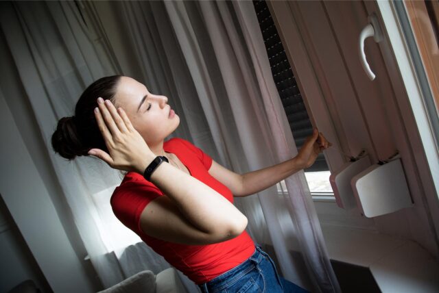 Eine Frau steht in ihrer Wohnung und schließt gerade einen Rolladen, um ihre Wohnung vor Hitze zu schützen. Ihr ist offensichtlich warm und sie hält sich vor Erschöpfung durch die Hitze die rechte Hand an ihre Schläfe.