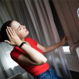 Eine Frau steht in ihrer Wohnung und schließt gerade einen Rolladen, um ihre Wohnung vor Hitze zu schützen. Ihr ist offensichtlich warm und sie hält sich vor Erschöpfung durch die Hitze die rechte Hand an ihre Schläfe.