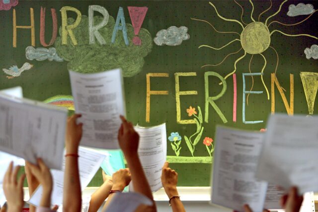Vor einer Tafel mit der Aufschrift „Hurra! Ferien“ halten Schülerinnen und Schüler Zeugnisse in die Höhe. Die Tafel ist mit Zeichnungen wie einer Sonne, Wolken und Blumen verziert.