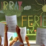 Vor einer Tafel mit der Aufschrift „Hurra! Ferien“ halten Schülerinnen und Schüler Zeugnisse in die Höhe. Die Tafel ist mit Zeichnungen wie einer Sonne, Wolken und Blumen verziert.