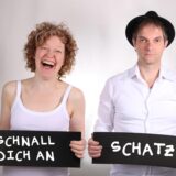 Zwei Personen posieren vor einem hellen Hintergrund und halten schwarze Schilder in den Händen. Die Frau auf der linken Seite lacht und trägt ein weisses Unterhemd, während der Mann rechts einen Hut trägt und ein weisses Hemd angezogen hat.