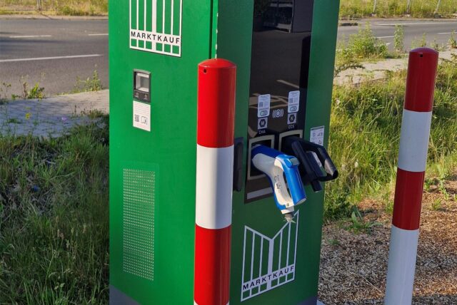 Eine grüne Ladesäule steht am Rande eines Parkplatzes. Die Kabel fehlen, nur noch die Stecker sind übrig.