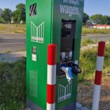 Eine grüne Ladesäule steht am Rande eines Parkplatzes. Die Kabel fehlen, nur noch die Stecker sind übrig.