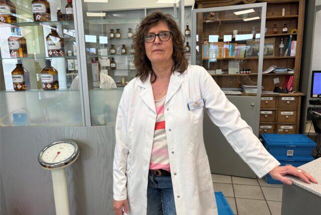 Eine Apothekerin, steht in der Apotheke, die sich durch ein historisches Interieur mit alten Apothekerflaschen und Schubladen auszeichnet. Im Hintergrund sind alte Gefäße mit Etiketten wie „Ethanol“ und „Benzin“ zu sehen.