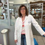 Eine Apothekerin, steht in der Apotheke, die sich durch ein historisches Interieur mit alten Apothekerflaschen und Schubladen auszeichnet. Im Hintergrund sind alte Gefäße mit Etiketten wie „Ethanol“ und „Benzin“ zu sehen.