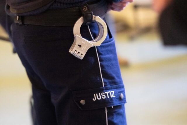 Ein Justizbeamter steht mit Handschellen im Verhandlungssaal eines Verwaltungsgerichts.