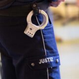 Ein Justizbeamter steht mit Handschellen im Verhandlungssaal eines Verwaltungsgerichts.