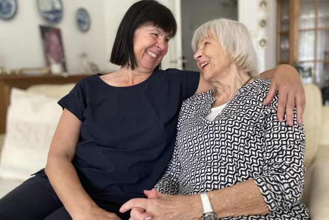 Zwei Frauen sitzen auf einem Sofa und lachen sich an. Eine der Frauen hat dunkles Haar, die andere graues.