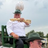 Eine Gruppe von Männern in Tracht trägt einen Mann auf einem Stuhl. Der Sitzende trägt ebenfalls eine Tracht mit einem Hut mit Federbusch und hält ein Stück Holz in den Händen.