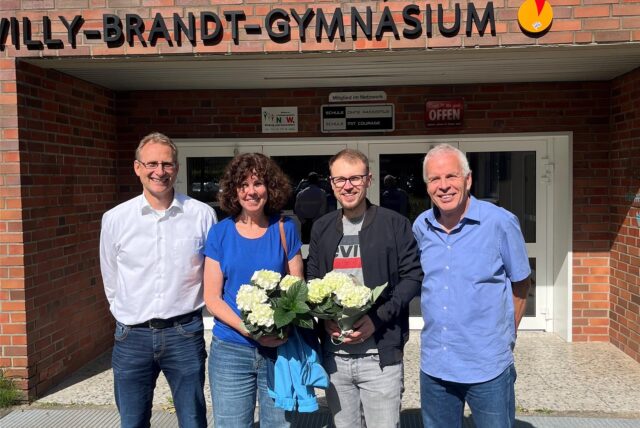 Vor dem Willy-Brandt-Gymnasium posieren vier Personen mit Blumensträußen vor dem Eingang des Gebäudes. Das Gymnasium engagiert sich als „Schule ohne Rassismus – Schule mit Courage“.
