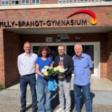 Vor dem Willy-Brandt-Gymnasium posieren vier Personen mit Blumensträußen vor dem Eingang des Gebäudes. Das Gymnasium engagiert sich als „Schule ohne Rassismus – Schule mit Courage“.