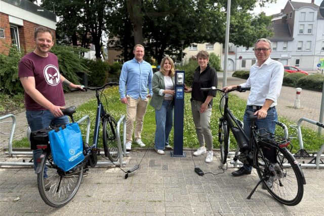Eine Gruppe von fünf Personen steht vor einer Ladesäule für Fahrräder im Freien. Zwei Fahrräder, ein blaues und ein schwarzes E-Bike, sind an einem Fahrradständer abgestellt und mit Ladegeräten an die Ladesäule angeschlossen.