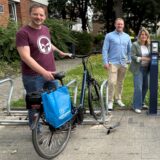 Eine Gruppe von fünf Personen steht vor einer Ladesäule für Fahrräder im Freien. Zwei Fahrräder, ein blaues und ein schwarzes E-Bike, sind an einem Fahrradständer abgestellt und mit Ladegeräten an die Ladesäule angeschlossen.