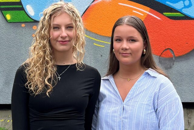 Nele (l.) und Lisa posieren vor einer Wand mit einem farbenfrohen Graffito, das den Namen der Schule (ASGSG) zeigt.