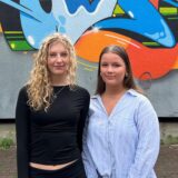 Nele (l.) und Lisa posieren vor einer Wand mit einem farbenfrohen Graffito, das den Namen der Schule (ASGSG) zeigt.