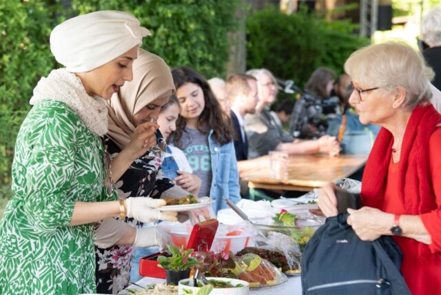Eine Frau mit Kopftuch verkauft Essen an einem Stand. Eine Besucherin mit grauen Haaren holt ihre Geldbörse aus der Tasche.