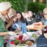 Eine Frau mit Kopftuch verkauft Essen an einem Stand. Eine Besucherin mit grauen Haaren holt ihre Geldbörse aus der Tasche.